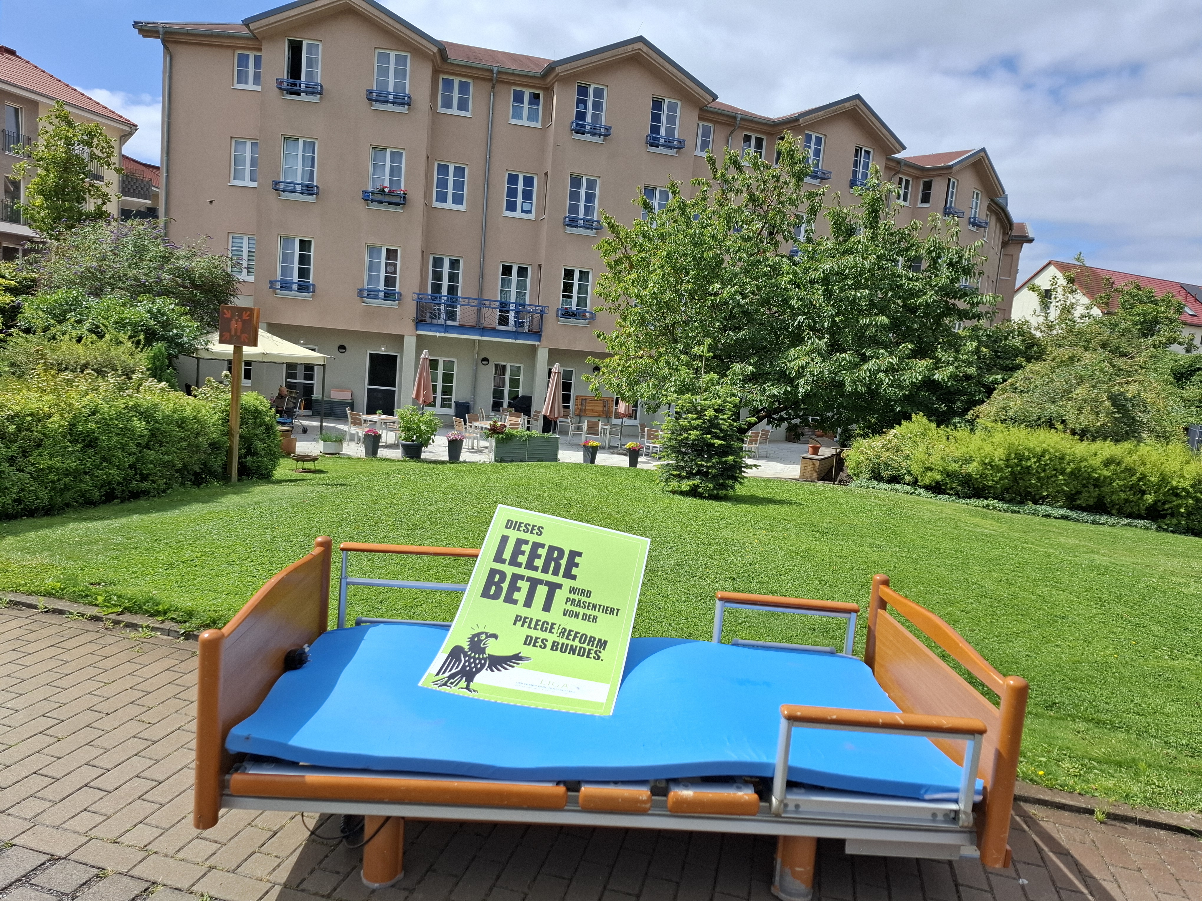 Leeres Pflegebett vor Rasenfläche und Einrichtung. Auf dem Bett liegt ein grünes Schild: "Dieses leere Bett wird präsentiert von der Pflegereform des Bundes."