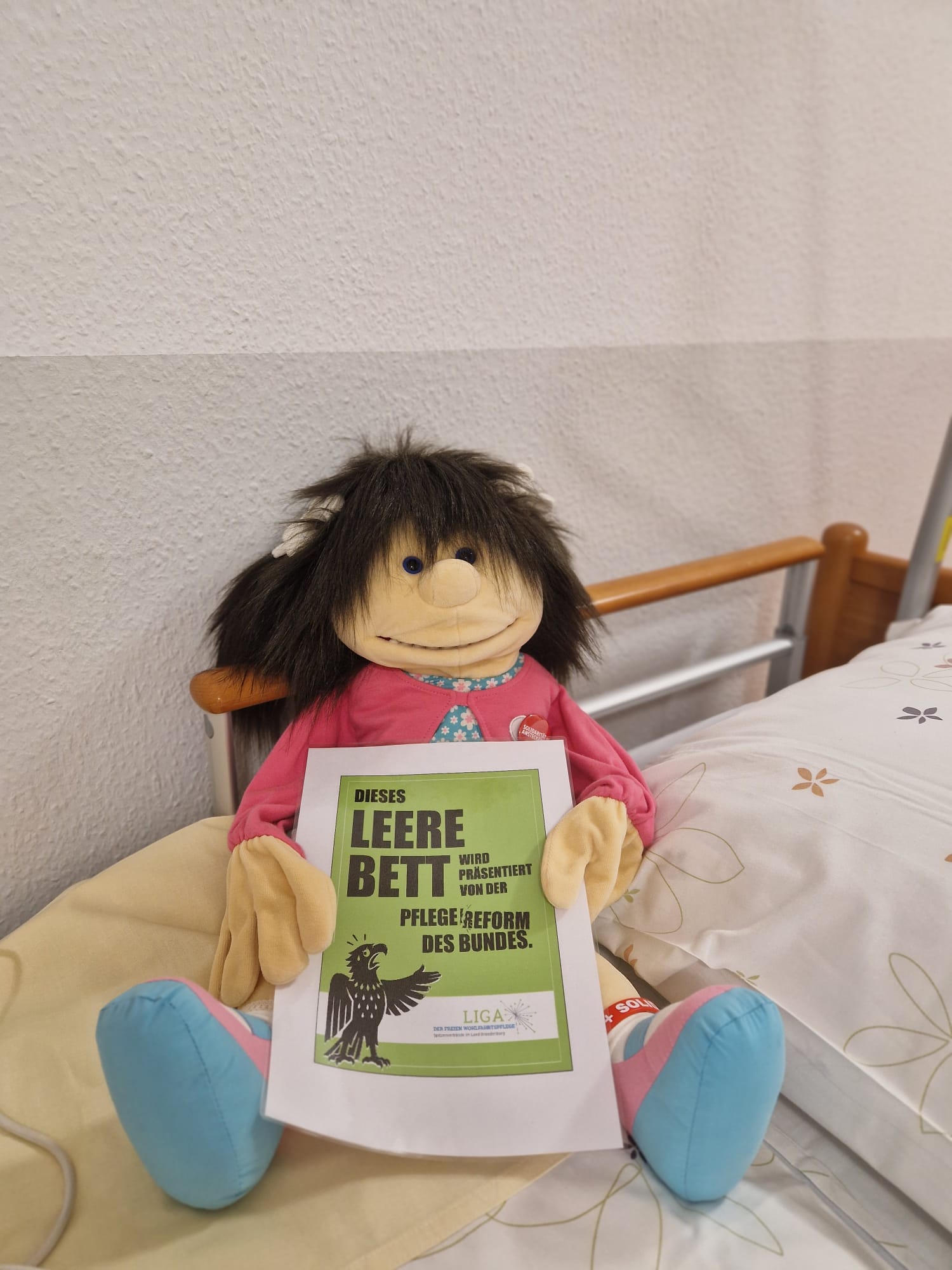 Plüsch-Puppr mit dunklen struppel Haaren sitzt im Pflegebett und hält Plakat DIN A4 grüner Hintergrund: Dieses leere Bett wird präsentiert von der Pflegereform des Bundes.