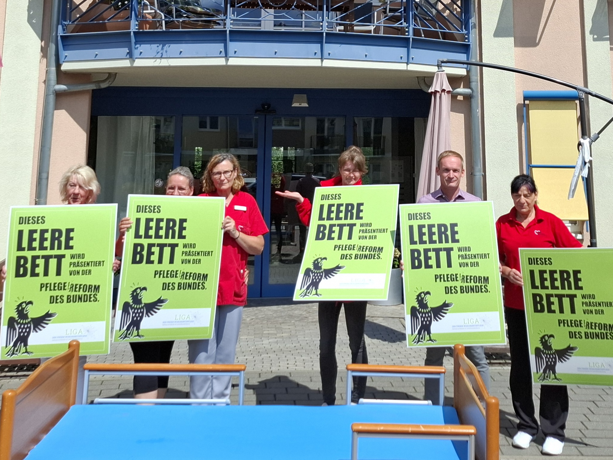 Ein leeres Pflegebett, dahinter 6 Pflegende der Einrichtung dahinter. Sie halten große grüne Schilder vor sich in die Kamera.