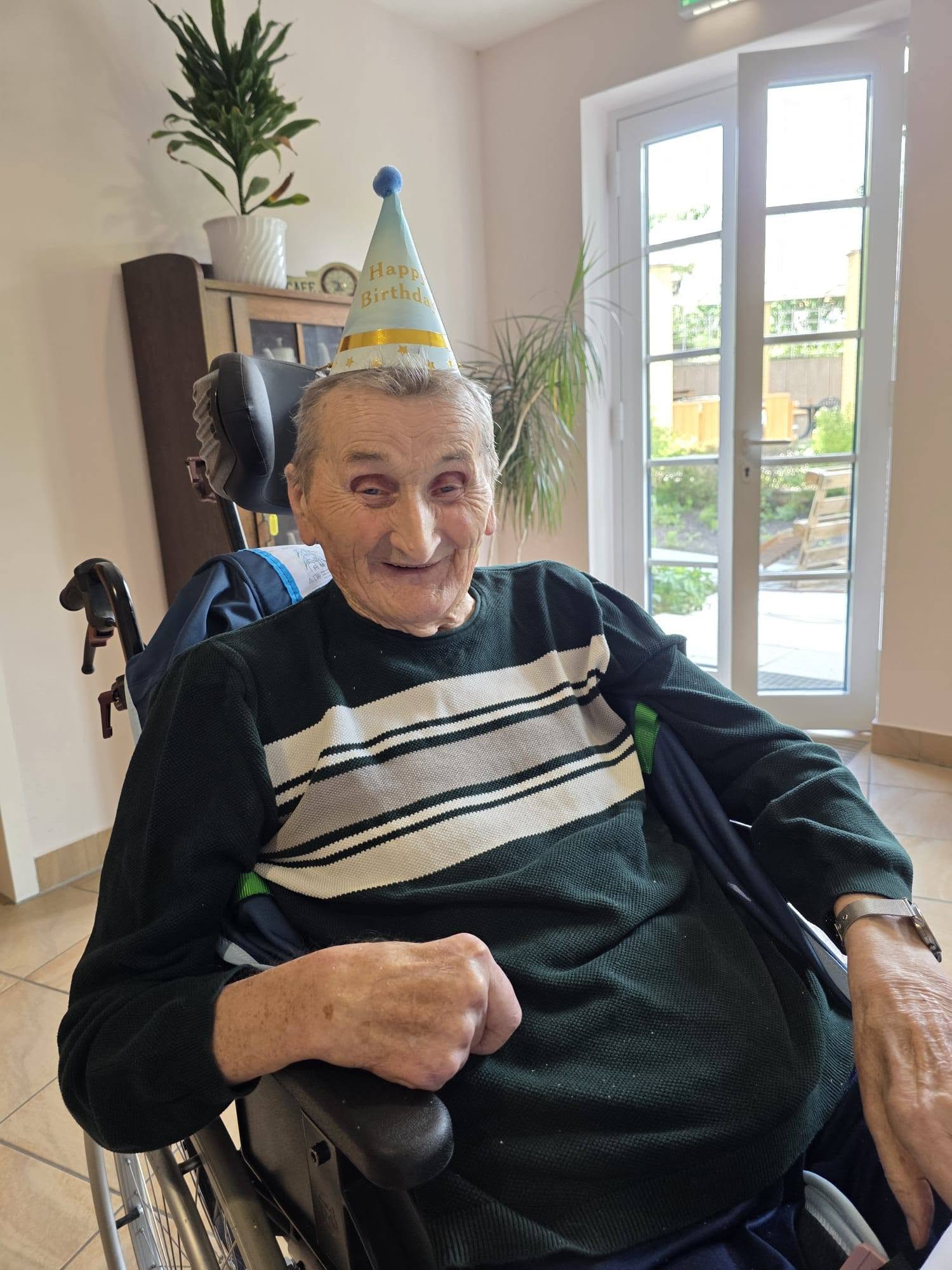 Sehr alter Mann im Rollstuhl mit Partyhütchen, er lächelt.