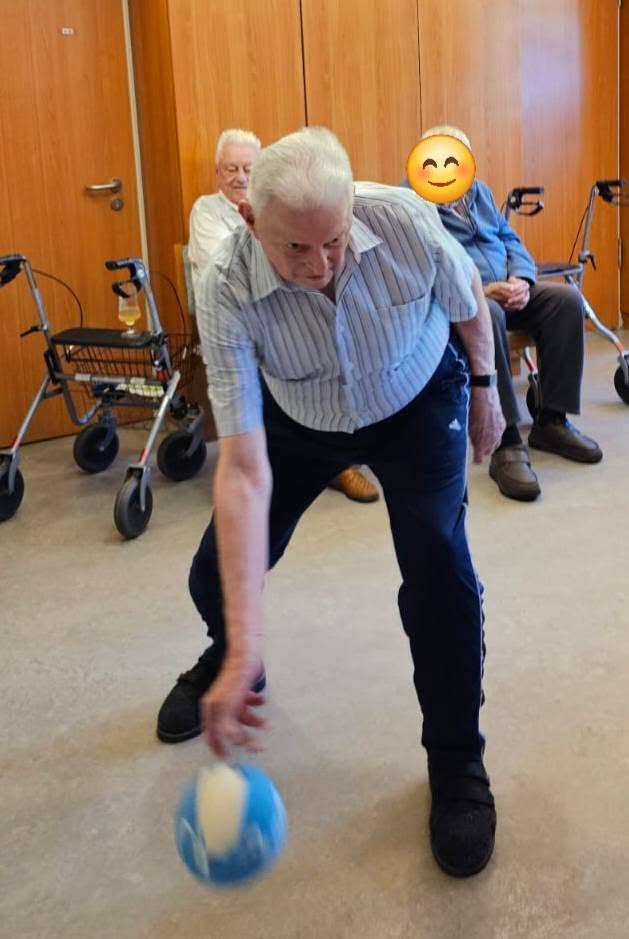 Ältere Mann wirft Ball am Boden.  Im Hintergrund sitzen zwei Personen.