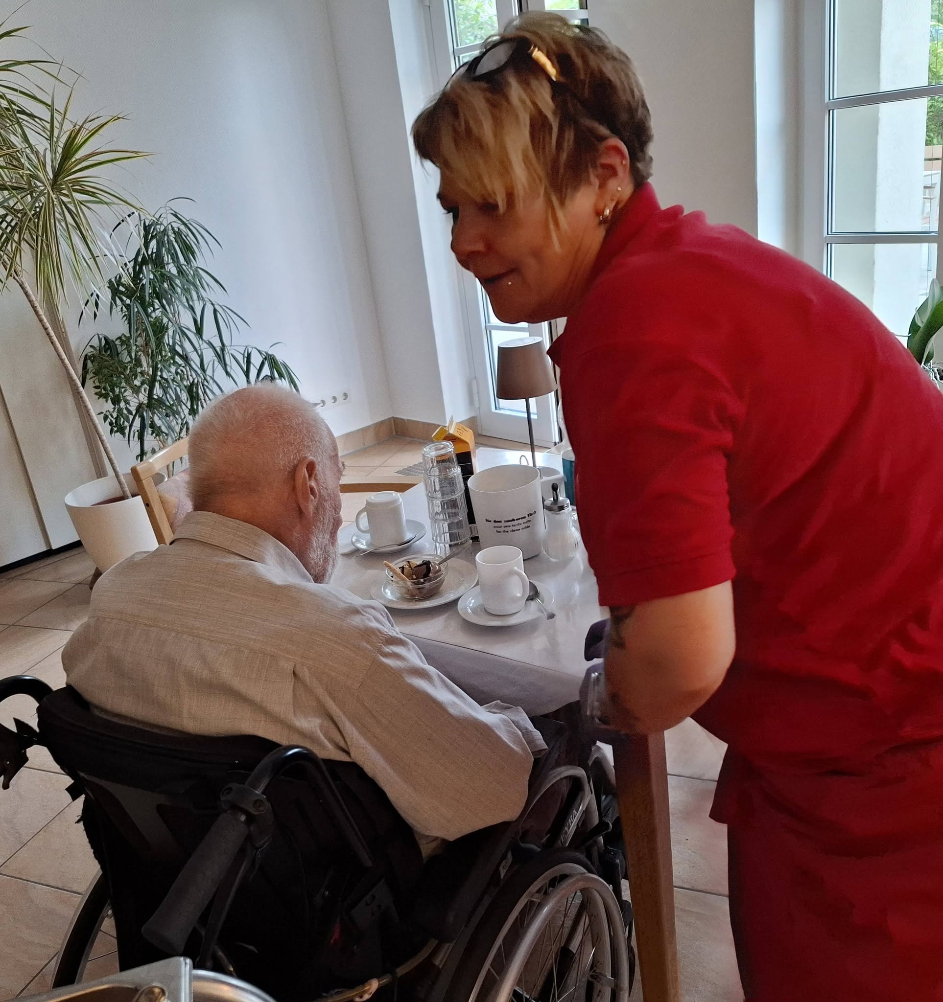 Senior am Tisch. Mit Eisbecheer und Kaffee vor sich. Pflegerin in roter Kleidung steht am Tisch.