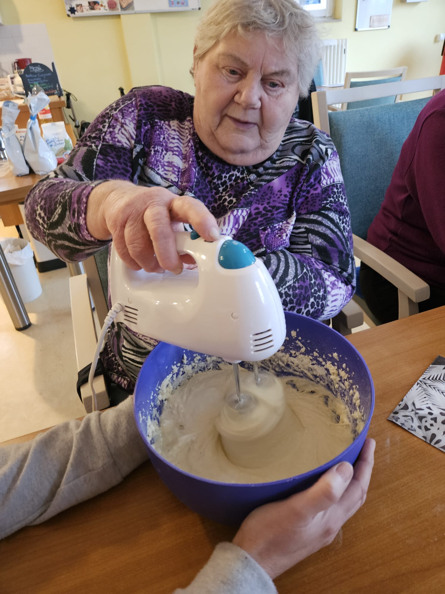 Seniorin mit Rührmaschine im Teig. Eine andere Person hält dabei die Schüssel fest.