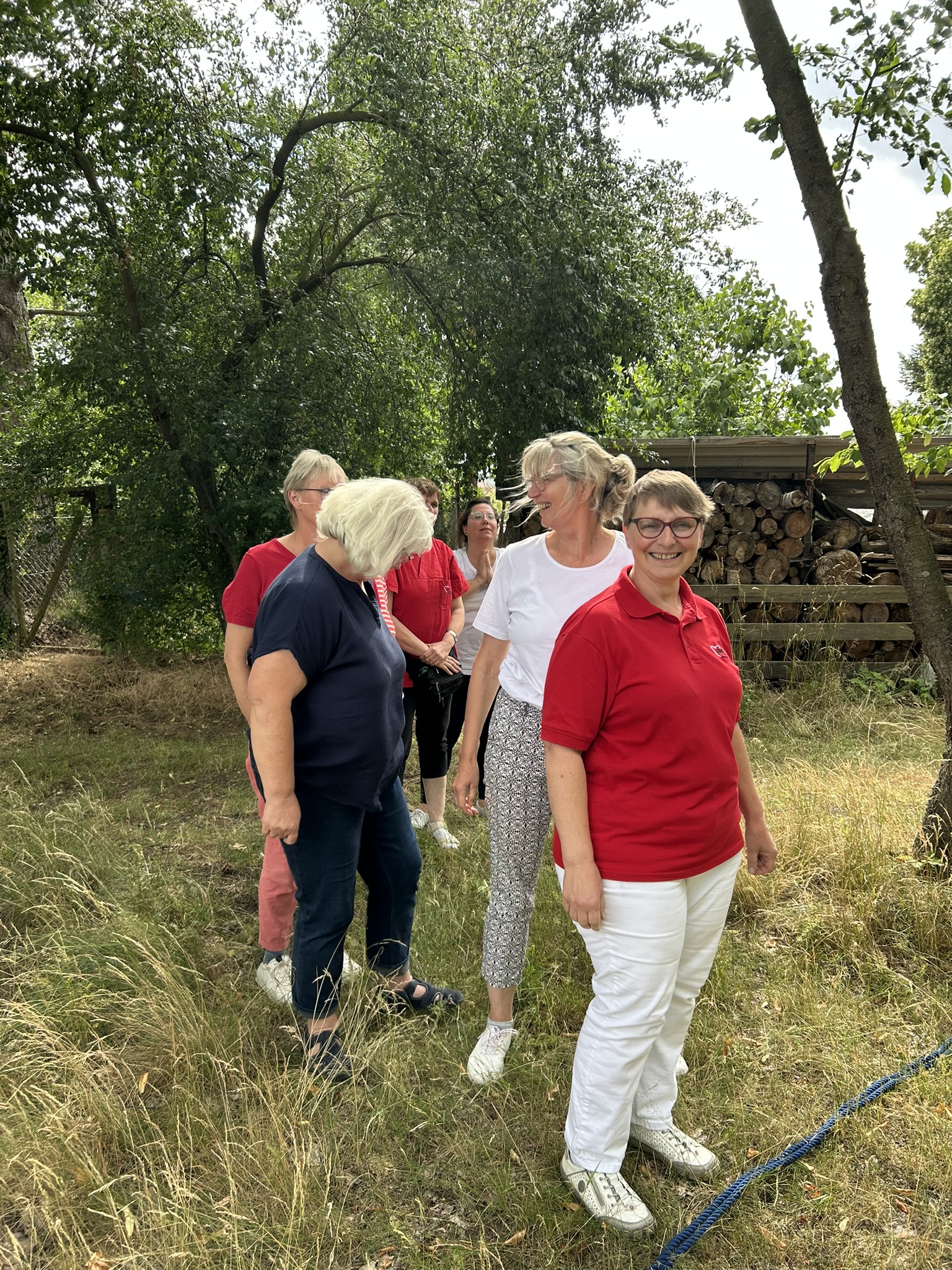 5 Personen stehen hintereinander aufgereit auf einer Wiese vor einem blauen Seil was unten auf dem Boden liegt, im Hintergrund befinden sich Bäume und gestapeltes Holz hinter einem Zaun