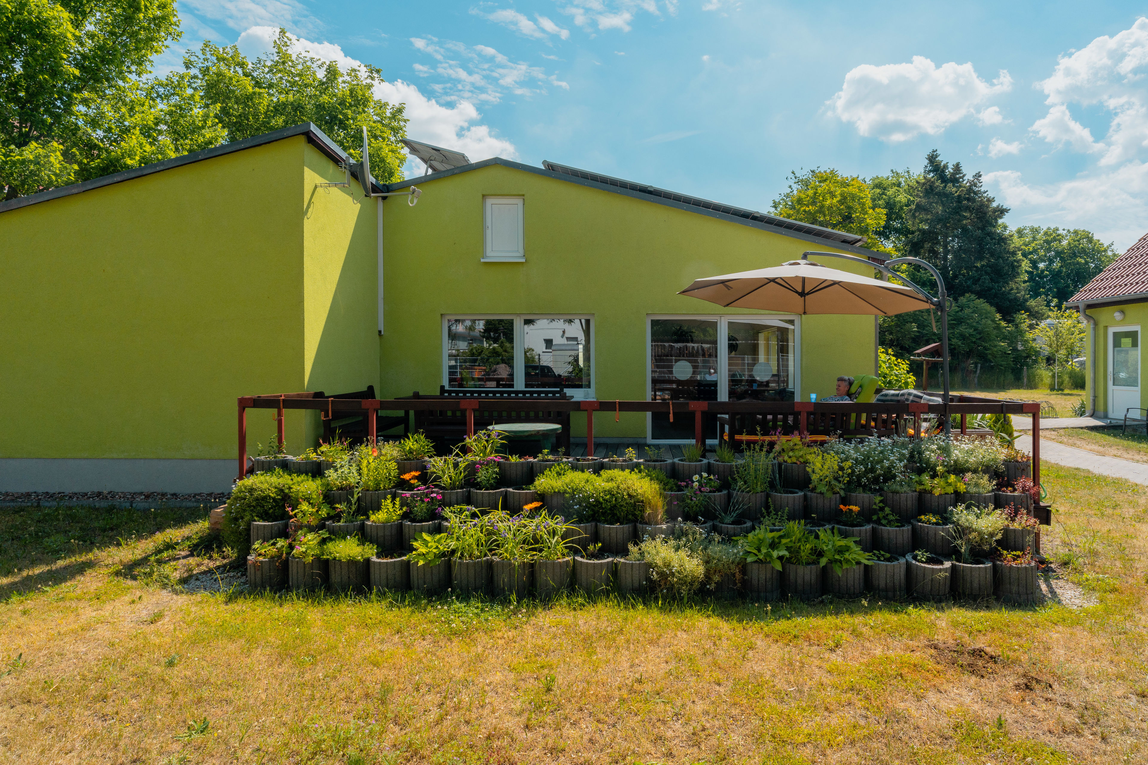 Seitenansicht von einem hellgrünen Haus mit einer Terrasse und Sonnenschirm. Viel Pflanzen davor.