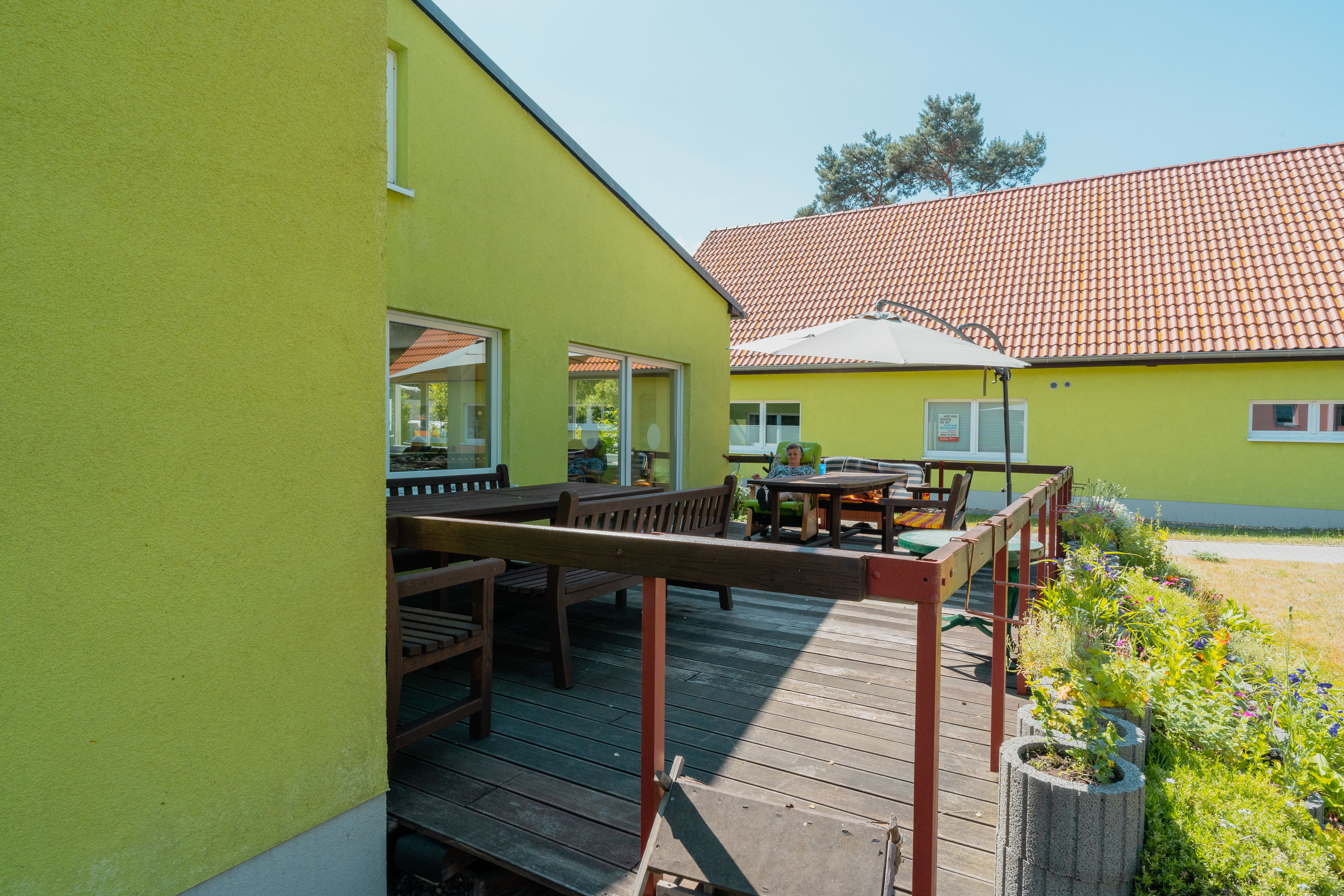 Terrasse mit Sonnenschirm, Tischen und Bänken eines hellgrünen Hauses.