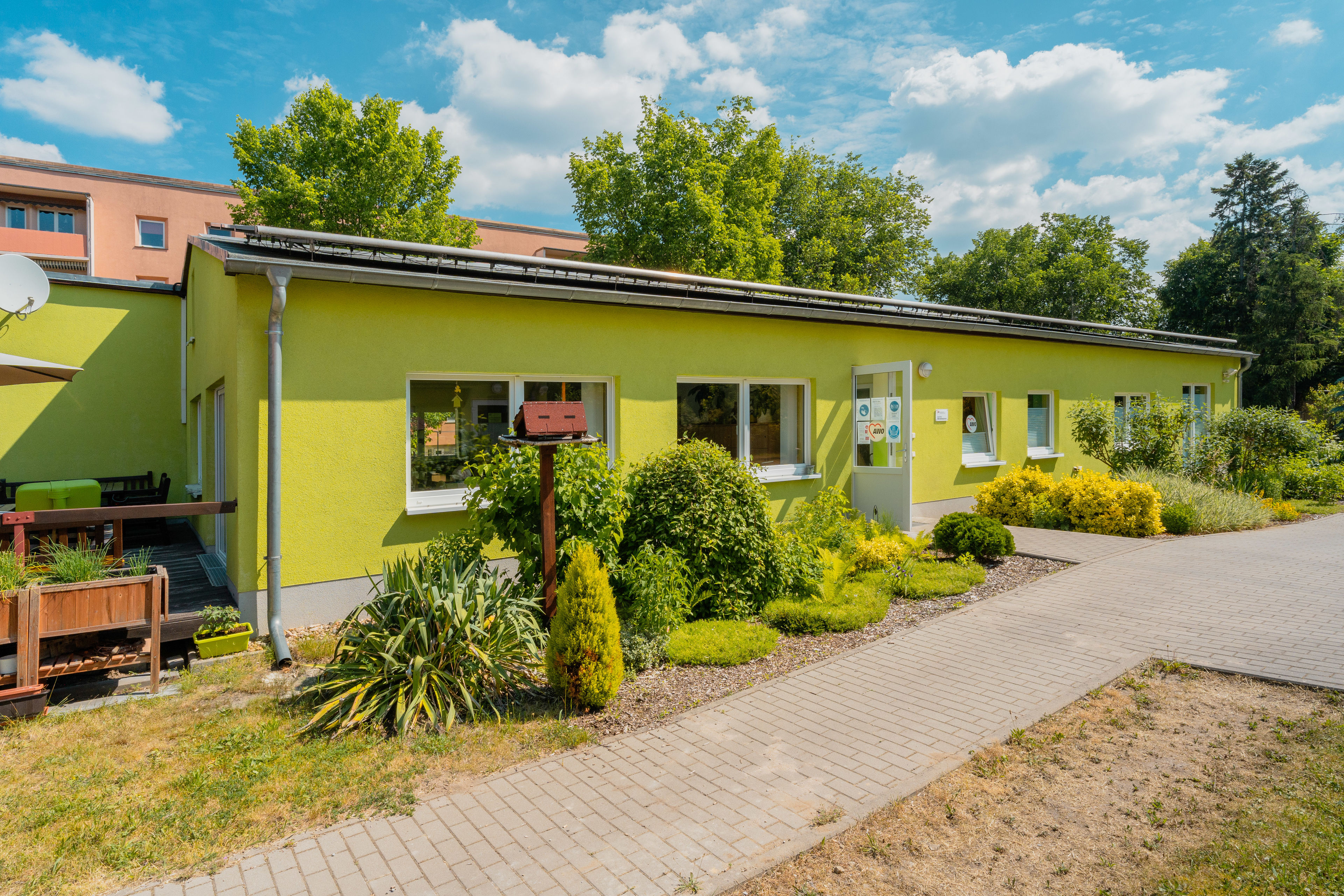 Grünes flaches Gebäude, davor Pflanzen und Büche, die Eingangstür steht offen.