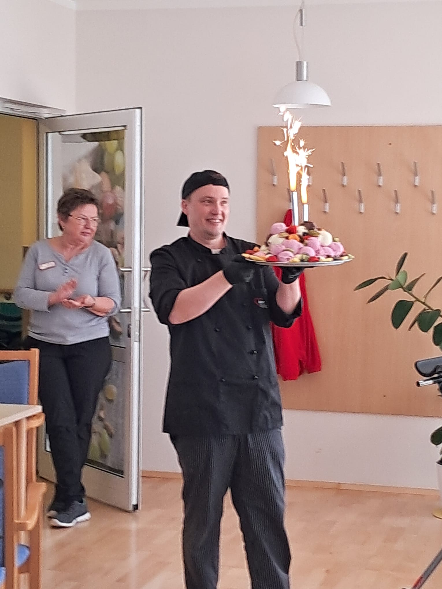 Küchenchef läuft bei den Frauen vorbei dabei hat auf einem Tablett bunte Eiskugeln gestapelt und zwei Leuchtfontänen verziehrt