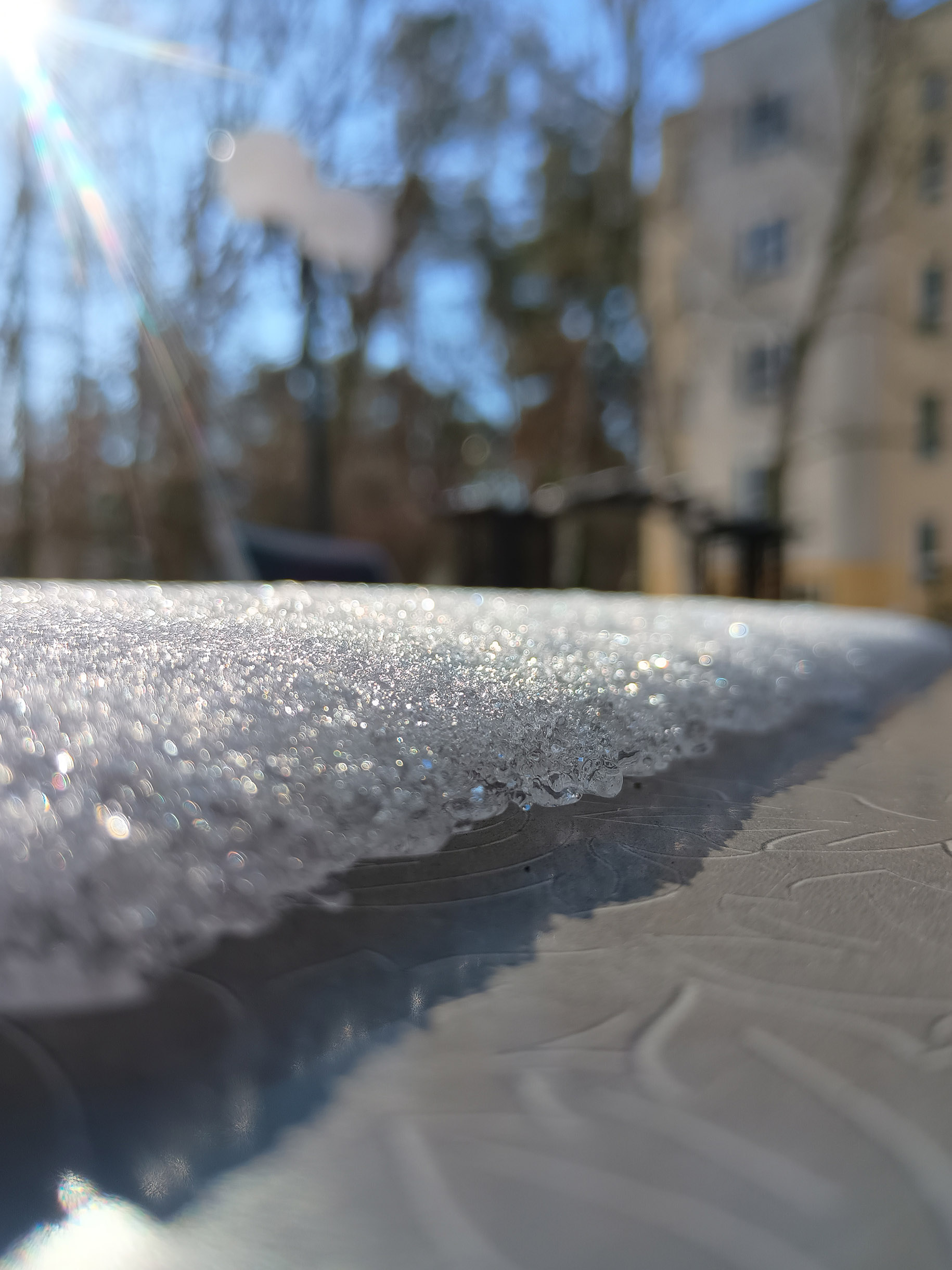 Schnee/Eis glitzert in der Sonne, im Hintergrund verschwommen Bäume und ein Haus.