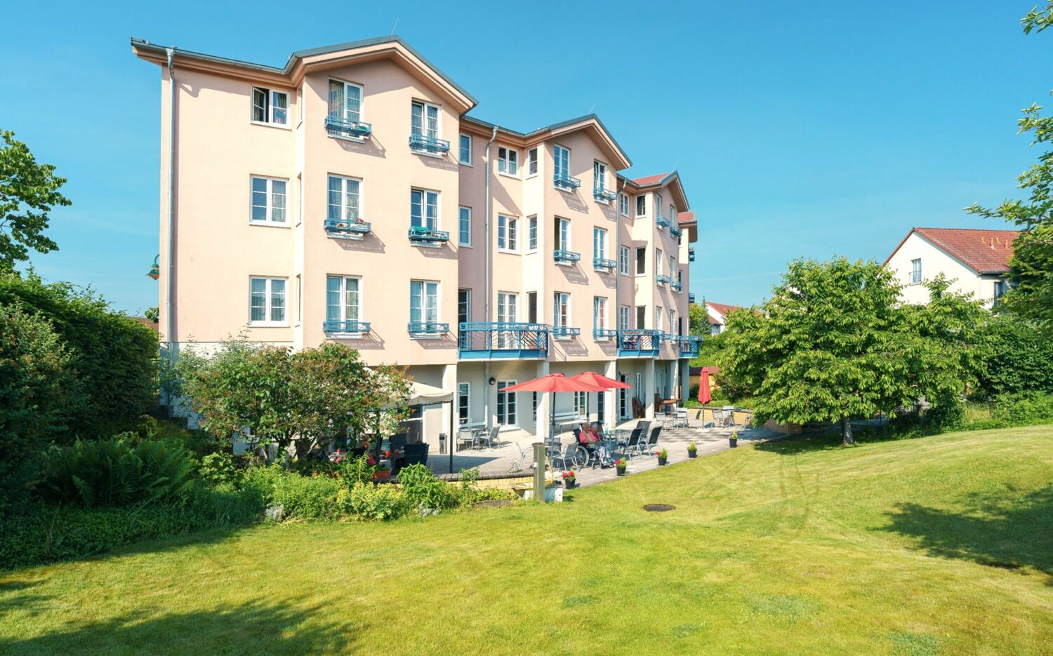 Großes Grundstück mit Rasen, Bäumen, Terrasse und vierstöckigen Gebäude mit Balkonen.