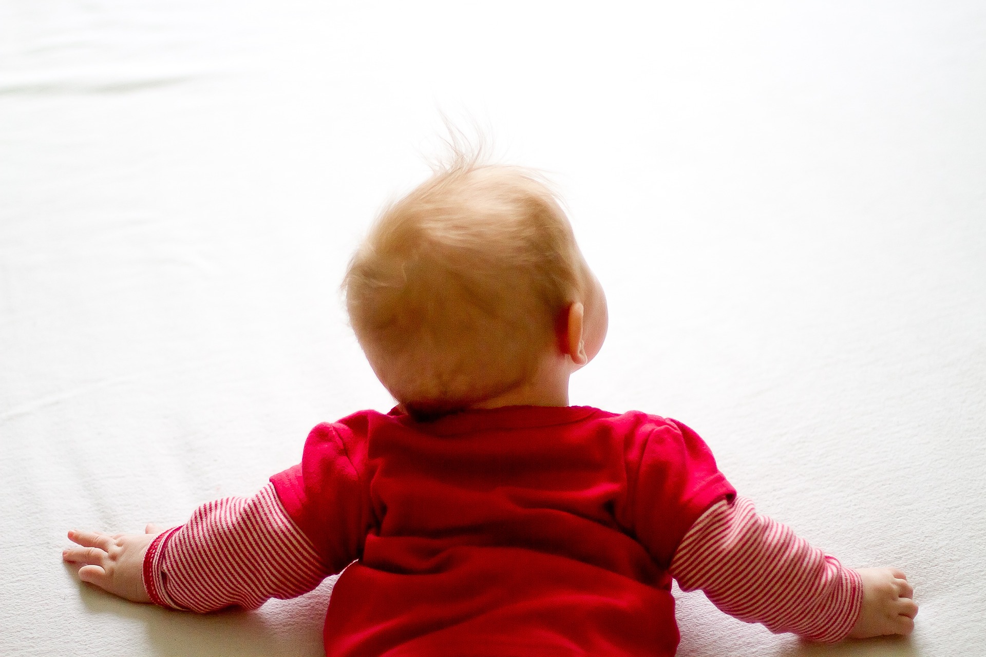 Ein Baby im roten Strampelanzug und hellen Haaren, welches auf dem Boden krabbelt. Von hinten oben.