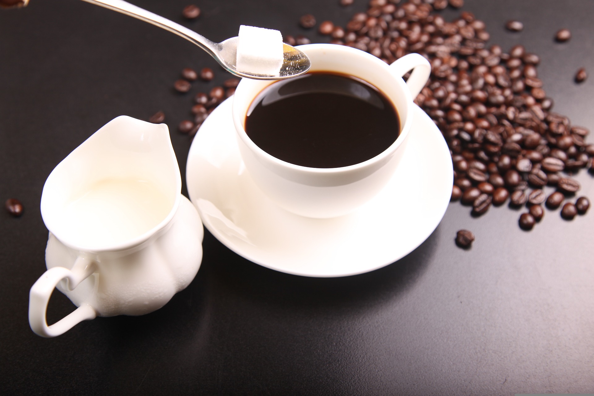 Eine weiße Tasse Kaffee. Ein weißes Kännchen mit Milch steht daneben. Mit einem Löffel wird ein Würfelzucker in den Kaffee gegeben. Auf dem Tisch sind Kaffeebohnen verstreut.