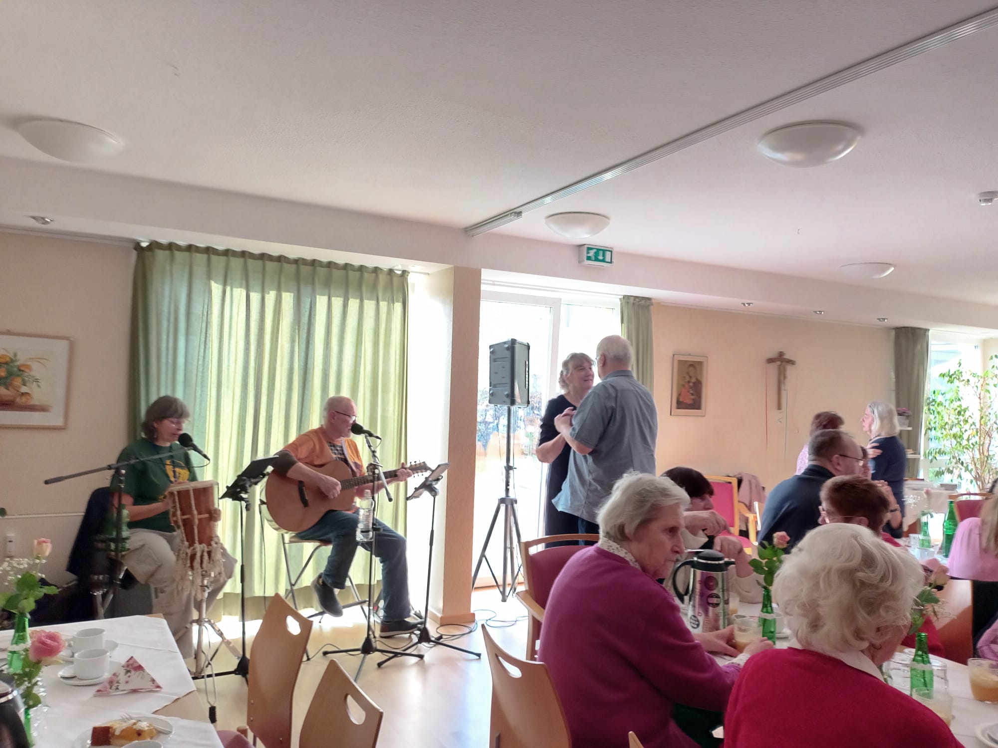 Heller Raum mit großen Fenstern. Kaffeetafeln und zwei Menschen die am Fenster sitzen und Musik machen.