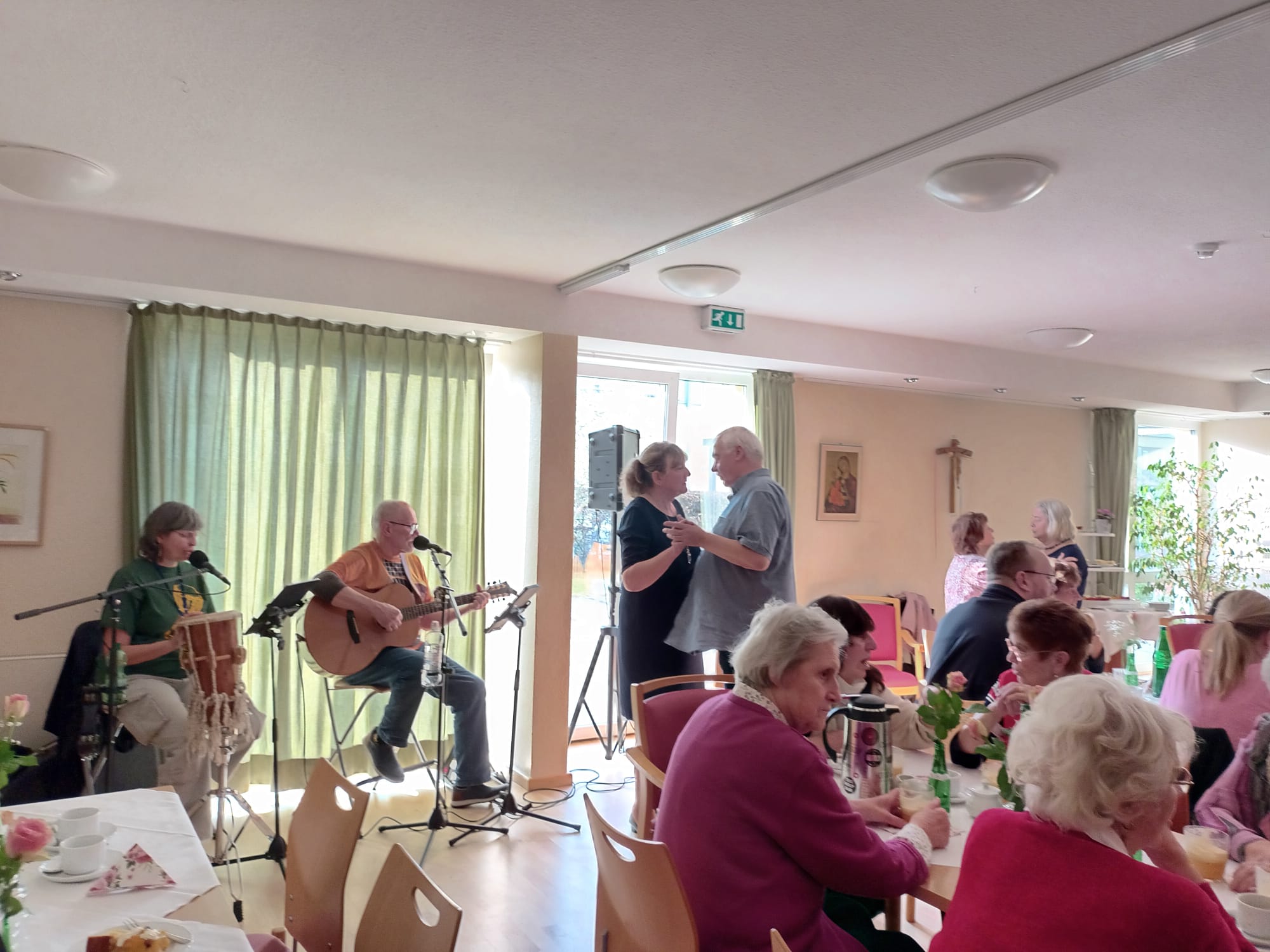 Heller Raum mit großen Fenstern. Kaffeetafeln und zwei Menschen die am Fenster sitzen und Musik machen. Rechts von dem Musikern ein tanzendes Paar.