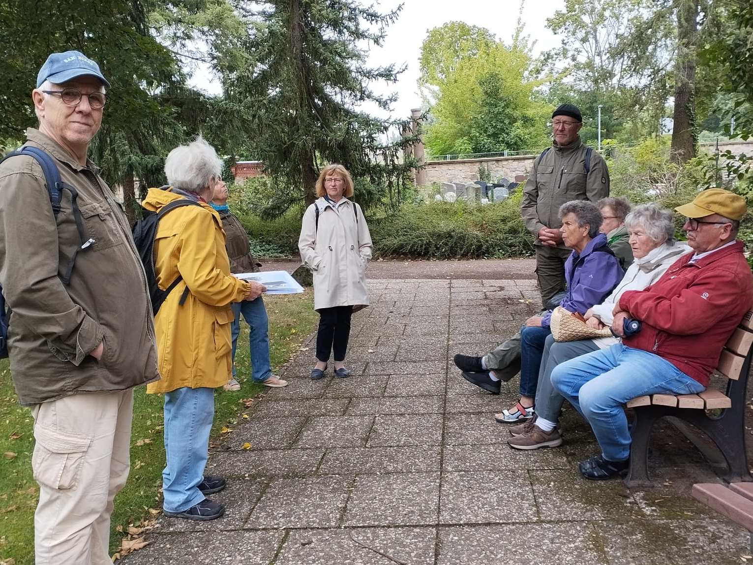 Links stehen Menschen in bequemer wetterfester Wanderkleidung und Rucksack. Links sitzen weitere Personen auf einer Bank.